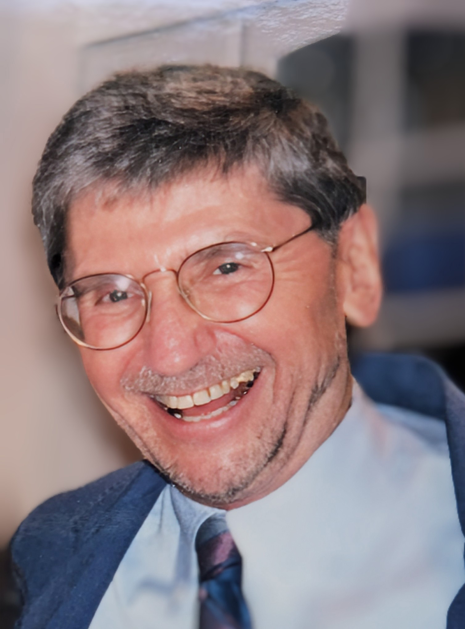 Richard Folke Gustafson smiles open-mouthed at the camera. His graying hair is cropped short and he has gray/black scruff on his face. He is wearing a dark blue blazer and a light blue button down with a blue and purple tie. He is wearing thin-rimmed circular glasses on his face. 